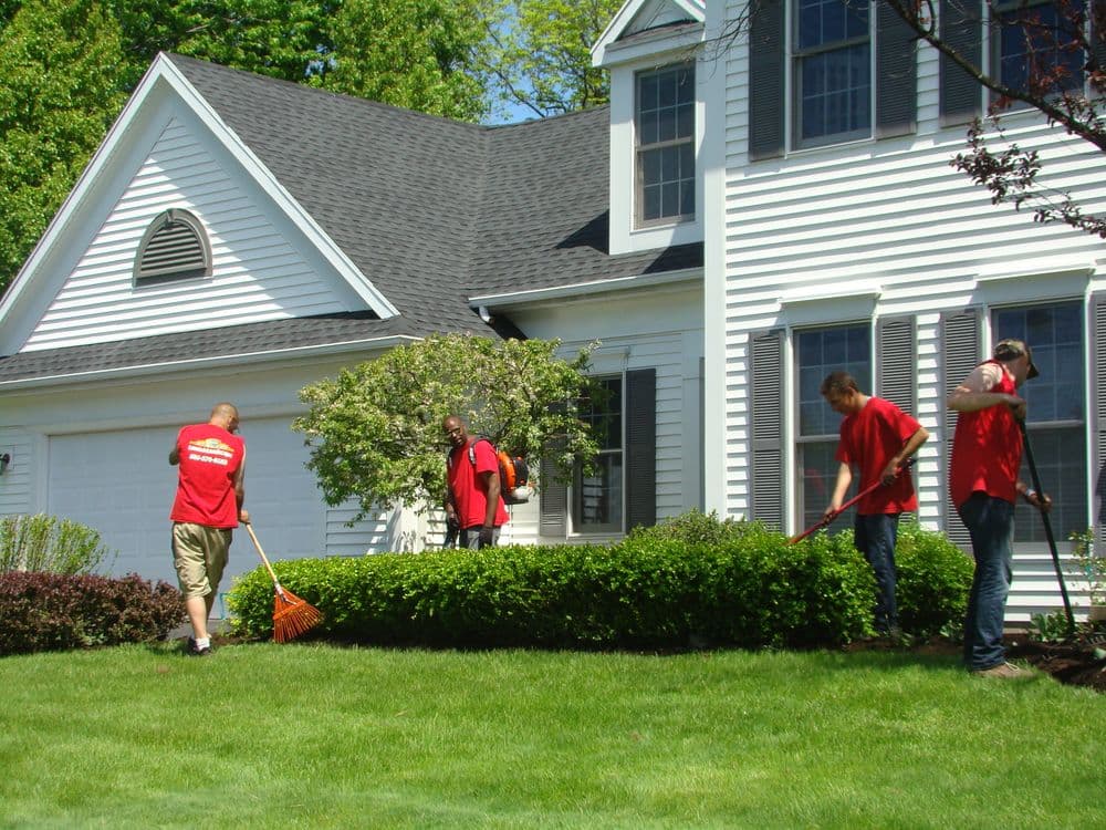 Landscapers in red shirts maintaining a green lawn and hedges near a home.