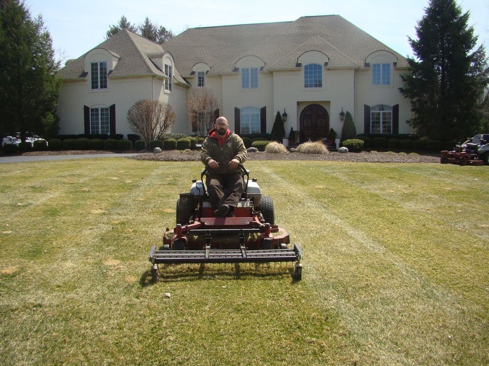 Man operating a zero-turn lawn mower on a well-manicured lawn in front of a large house.