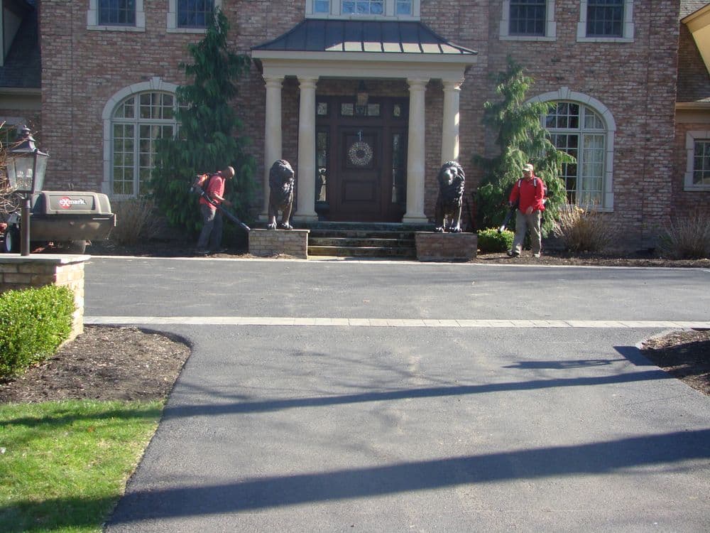 Landscapers maintaining driveway near brick mansion with lion statues at entrance.