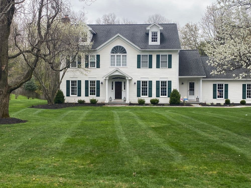 Colonial-style white house with green shutters and manicured lawn, surrounded by trees.