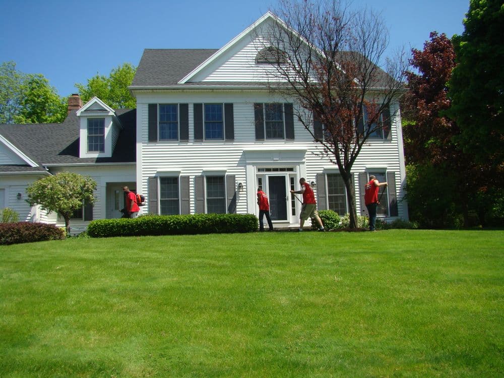 Two individuals in red jackets standing in front of a classic white house with a green lawn.