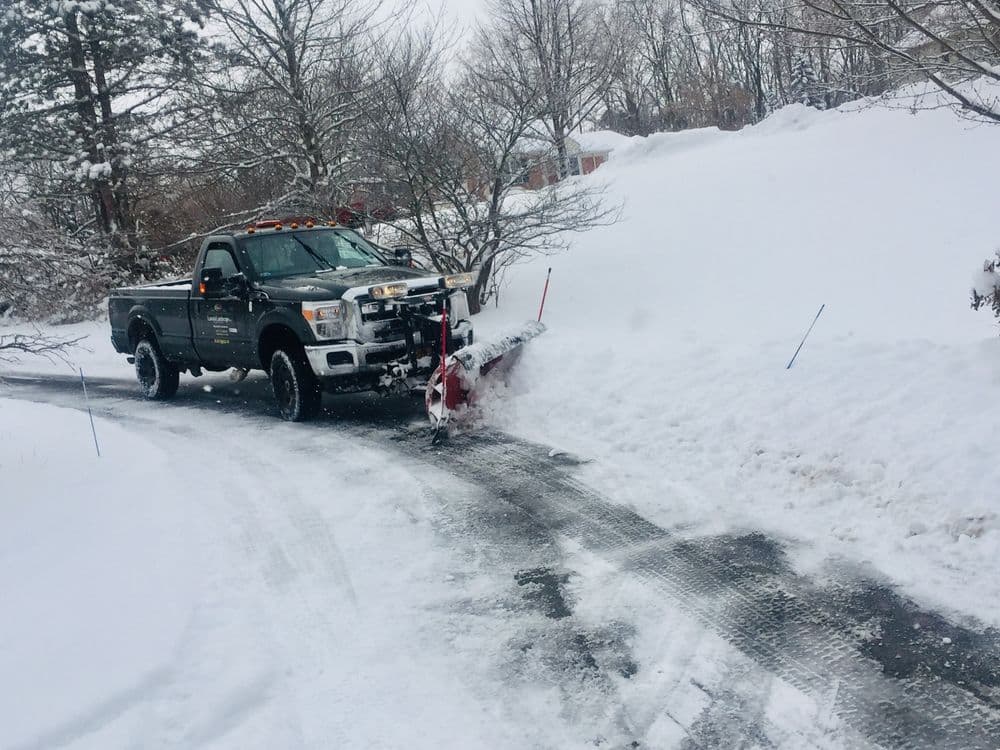 Snow plow truck clearing a residential driveway covered in heavy snow. Winter weather scene.
