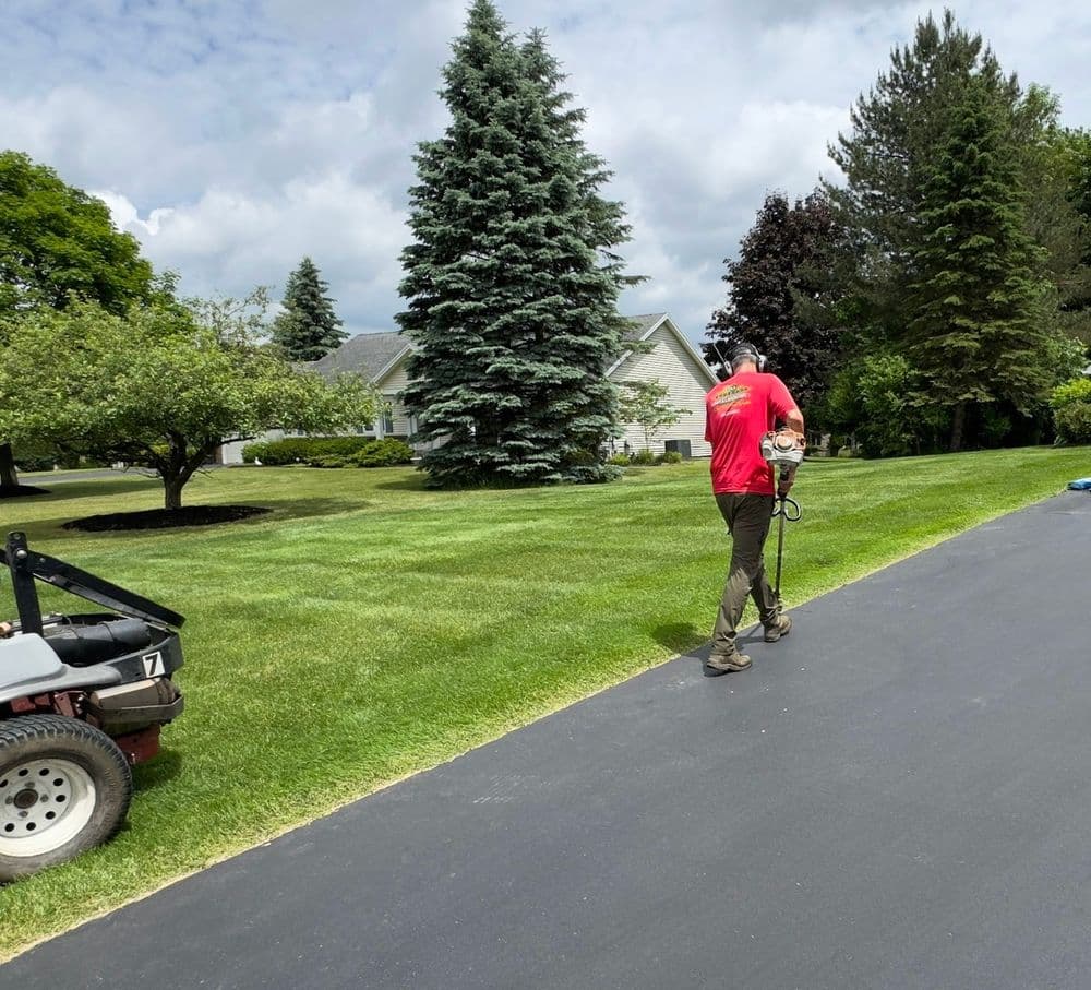 Worker in red shirt uses a lawn care tool on a well-maintained grassy lawn with trees.